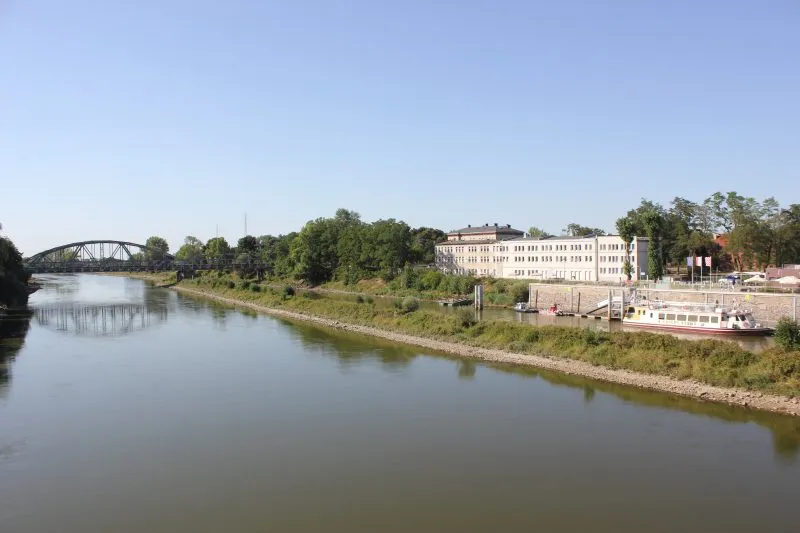 widok z góry na szeroką rzekę, po drugiej stronie rzeki znajduję się biały podłużny budynek w oddali widać brązowy most