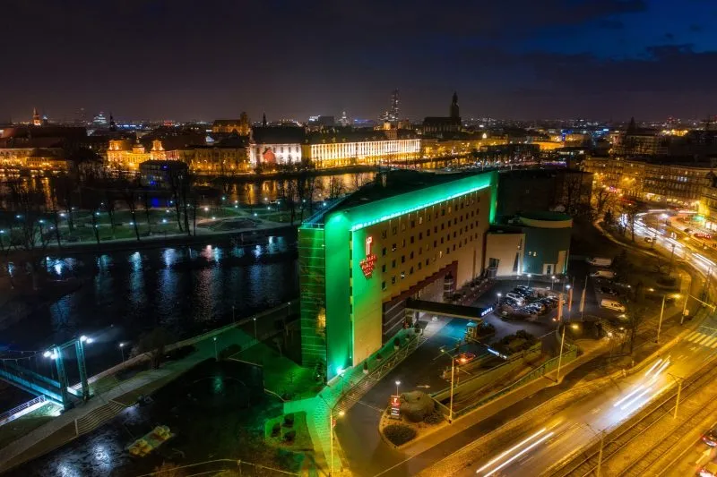 Widok na oświetlony budynek hotelu na tle rzeki oraz budynku Uniwersytety Wrocławskiego.