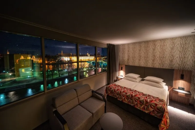 Pokój hotelowy z dużym oknem z widokiem na podświetlony gmach uniwersytetu wrocławskiego. W pokoju duże łóżko podójne, sofa oraz okrągły stolik.