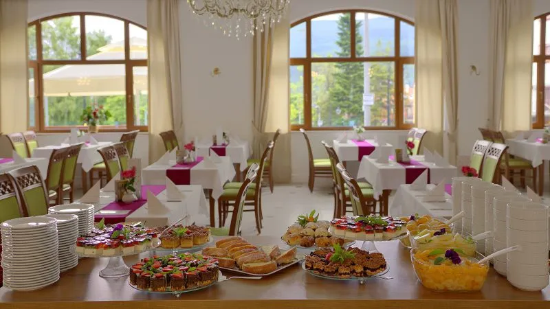 Przestronna, duża, jasna sala restauracyjna ze stołami nakrytymi białymi obrusami z bordowym bieżnikiem. Na pierwszym planie stół z deserami - ciasta, owoce w syropie, drobne wypieki.