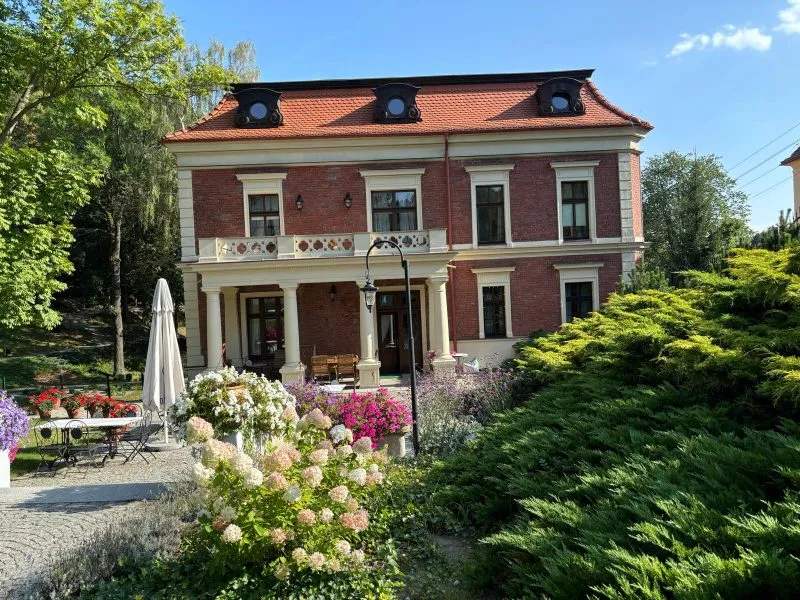 Odrestaurowany budynek eleganckiej willi z tarasem osadzonym na kolumnach na tle ogrodu pełnego zieleni. 