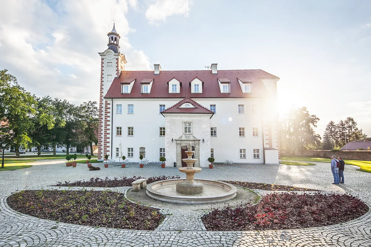 Prostokątny budynek pałacowy z wąską wysoką wieżą od strony północnej na tle zachodzącego słońca. Przed wejściem fontanna i o zdobny kolisty klomb.