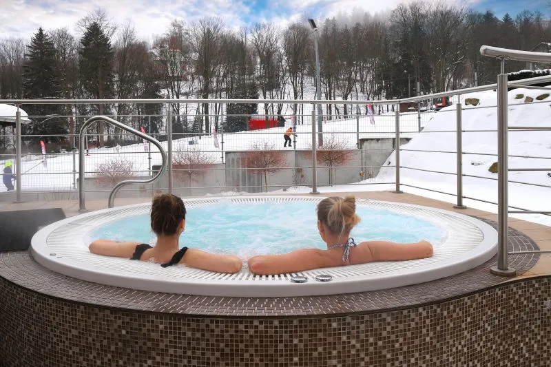Zewnętrze, okrągłe jacuzzi, w którym leżą dwie kobiety, wpatrzone w zimowy krajobraz.