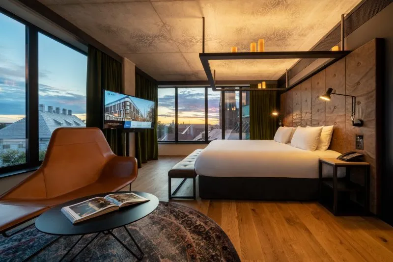Zdjęcie przedstawia nowoczesny, elegancki pokój hotelowy urządzony w szarej, minimalistycznej stylistyce. Dominują w nim kolory – czerń, szarość i drewno. W pokoju znajdują się dwa łóżko typu king size z drewnianym zagłówkiem. Dwie ściany zostały przeszklone, roztacza się z nich piękny widok na zabytkowe budynki Ostrowa Tumskiego.