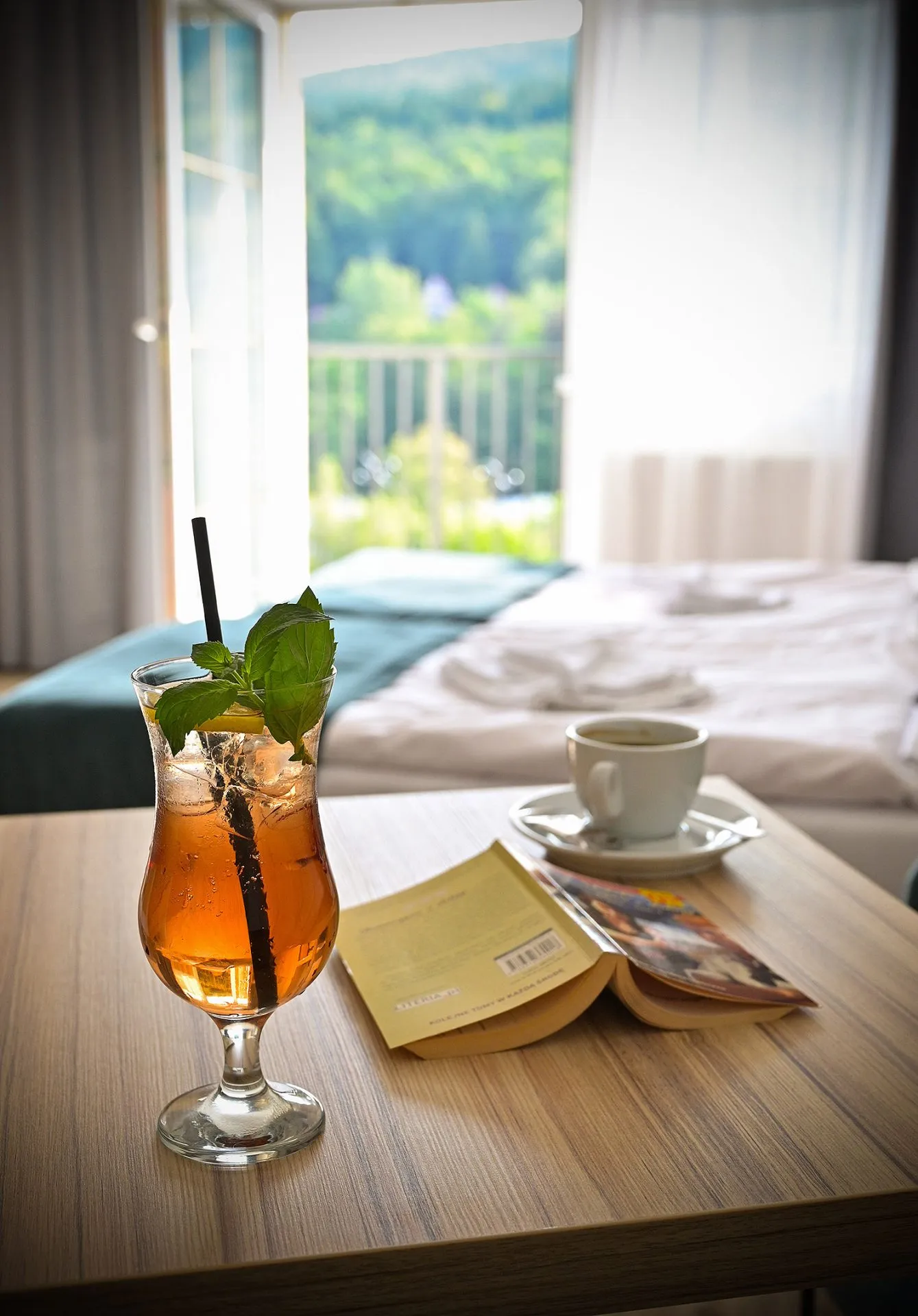 Elegancki drink ulokowany w towarzystwie książki i filiżanki z kawą na ławie w pokoju hotelowym z widokiem na las.