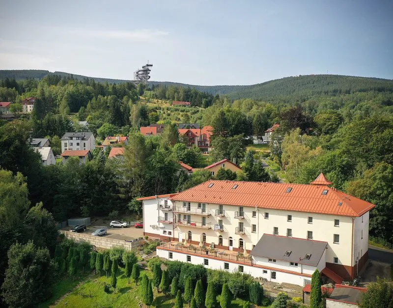 The extensive bright building of the Dobrzyński Resort with a red roof stands on a green slope, surrounded by a forest, a garden, and a regular row of tall shrubs.