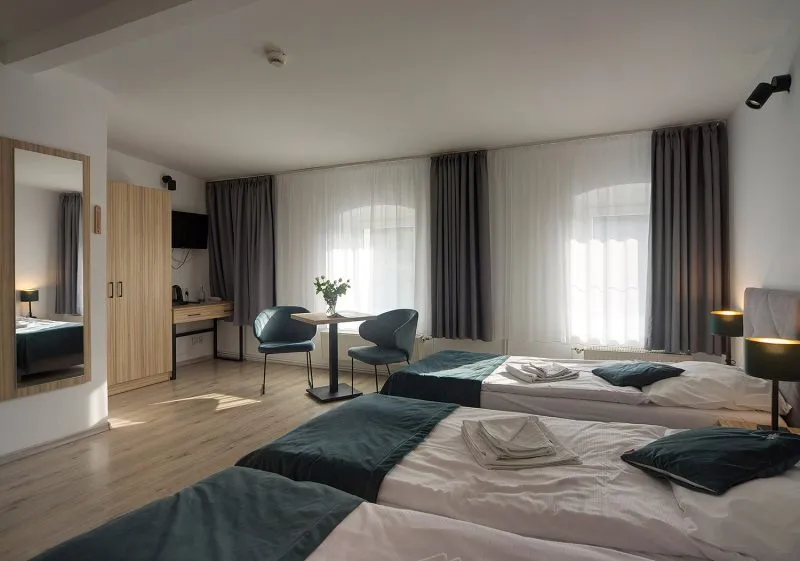 Pokój hotelowy z trzema łóżkami pojedynczymi, dużą szafą, telewizorem, stolikiem oraz dwoma fotelami.