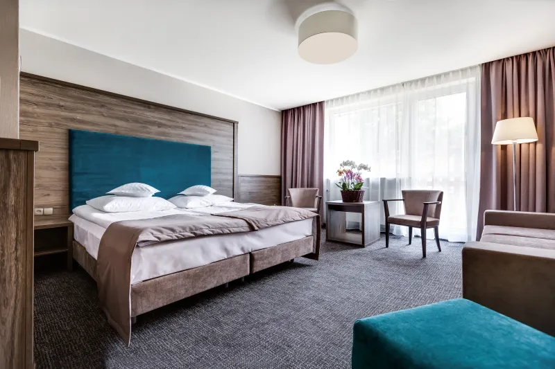 Elegancki pokój hotelowy z łóżkiem podwójnym. Nad łóżkiem drewniane obicie, na podłodze ciemna wykładzina. Pod dużym oknem, stolik z krzesłami.