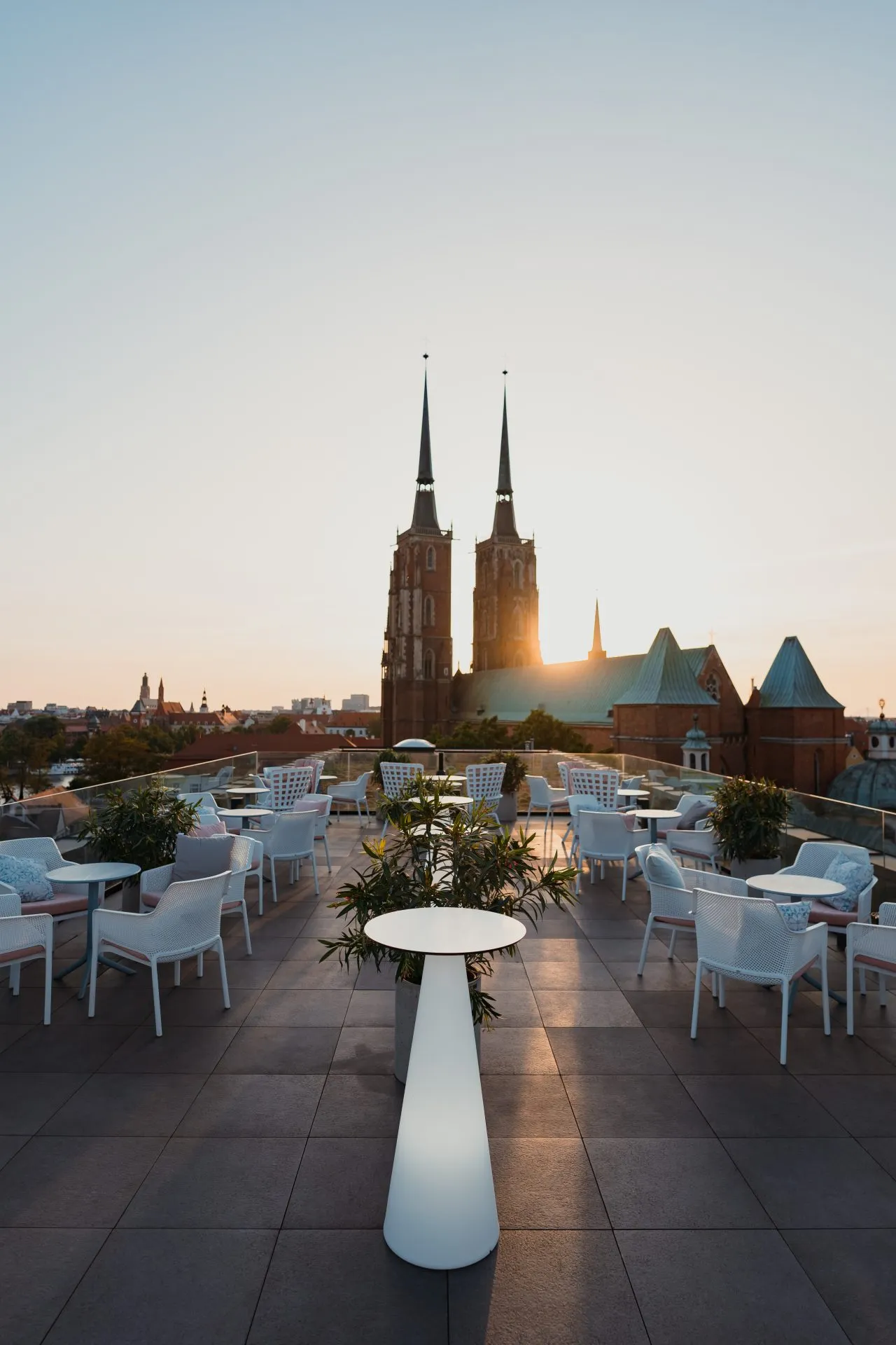 Zdjęcie przedstawia elegancki taras widokowy na dachu hotelu The Bridge Wrocław o zachodzie słońca. W centralnej części kadru widać nowoczesne białe stoliki i krzesła, a także dekoracyjną roślinność, która dodaje przestrzeni przytulności. W tle majestatycznie góruje Katedra Wrocławska.