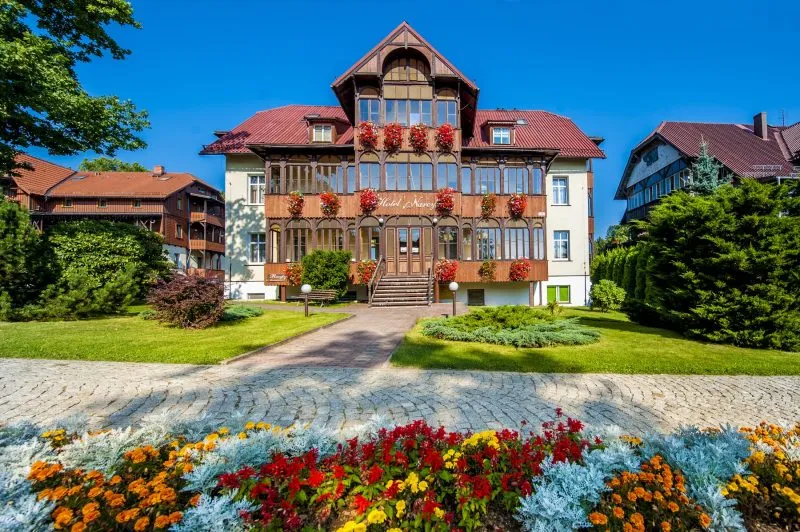 hotel od frontu, z dobrze widocznym wejściem i otaczającym go terenem. Jasna kolorystyka budynku i duże okna podkreślają jego elegancki, uzdrowiskowy styl.