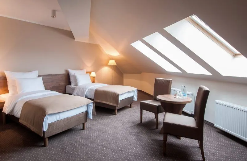 Pokój hotelowy, ulokowany na poddaszu z dwoma pojedynczymi łóżkami, okrągłym stolikiem i dwoma fotelami. Na podłodze wykładzina.