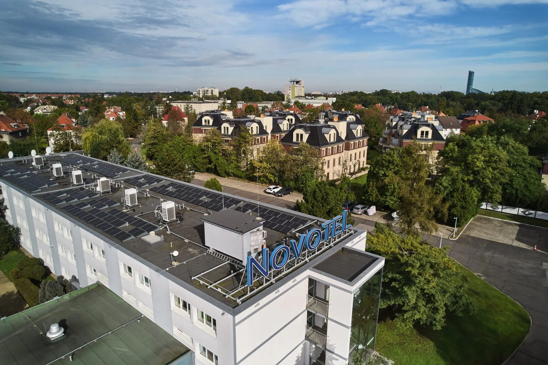 Widok z góry na budynek oznaczony niebieskim napisem „Novotel”, umieszczonym na dachu. W tle rozciągają się drzewa, osiedla i samotna wieża. Niebo jest lekko zachmurzone, nadając scenie spokojny nastrój. Hotel znajduje się pośrodku zielonego, miejskiego krajobrazu,