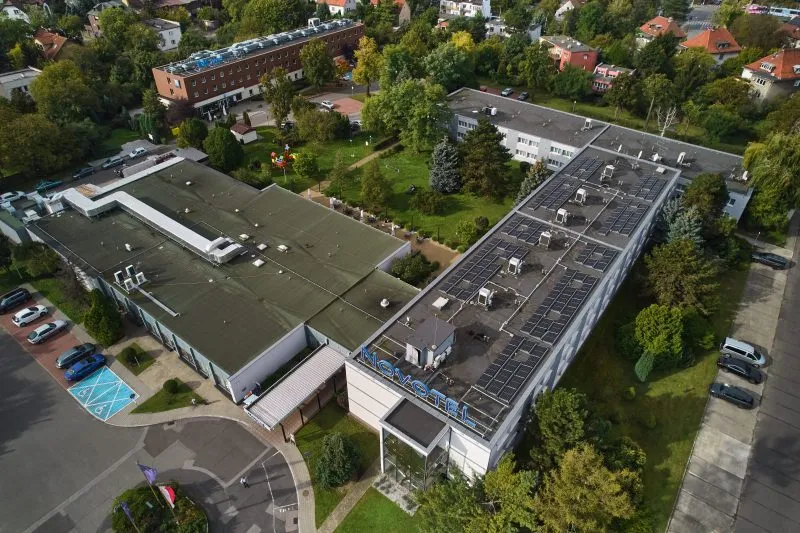 Zdjęcie ukazuje budynek hotelu otoczony zielenią. Przed hotelem znajdują się samochody oraz fragmenty trawnika. W tle widać zabudowania mieszkalne. Sceneria wygląda zadbanie i przyjaźnie.