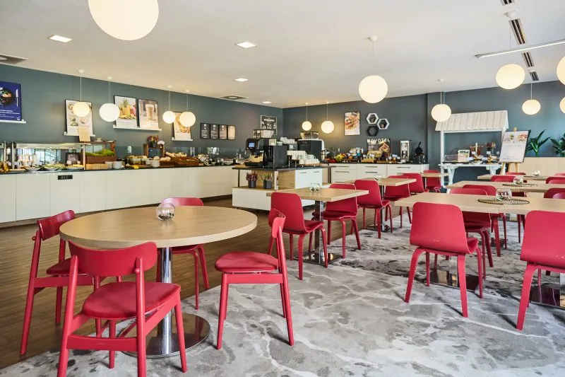 Zdjęcie przedstawia wnętrze nowoczesnej restauracji. W pomieszczeniu dominują czerwone krzesła, drewniane stoły i wiszące lampy sufitowe. W głębi znajduje się bar z butelkami i szklankami. Wystrój tworzy elegancki i przyjazny klimat.
