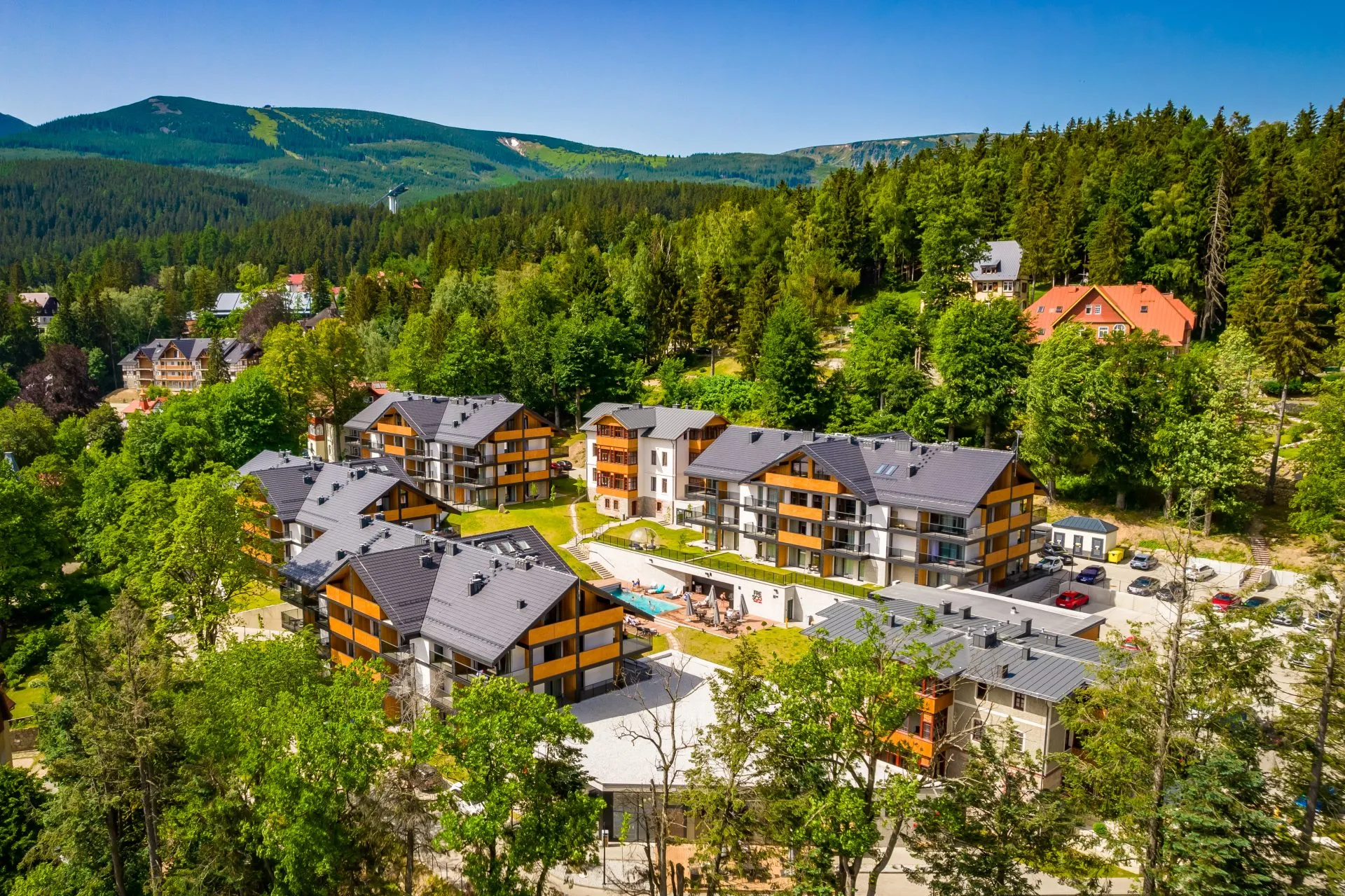 kompleks apartamentowy położony wśród zieleni u podnóża gór, otoczony lasem i górami w tle. Budynki mają drewniane akcenty elewacyjne, a w centralnej części znajduje się basen z leżakami.