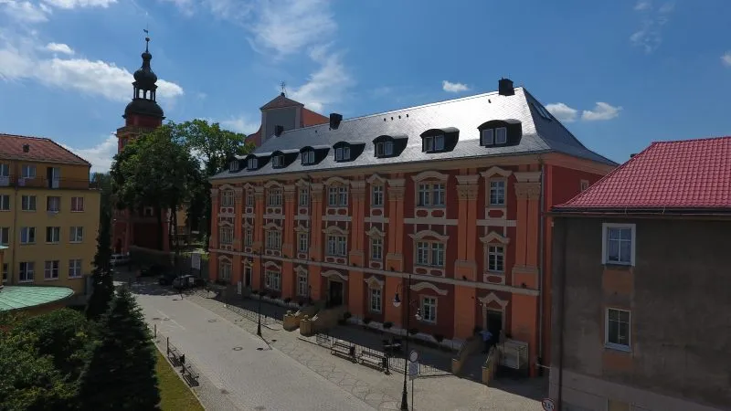 Długi, trójkondygnacyjny barokowy budynek, pięknie zrewitalizowany, pokryty kolorami w odcieniach pomarańczy i czerwieni. Na drugim planie wieża kościelna.