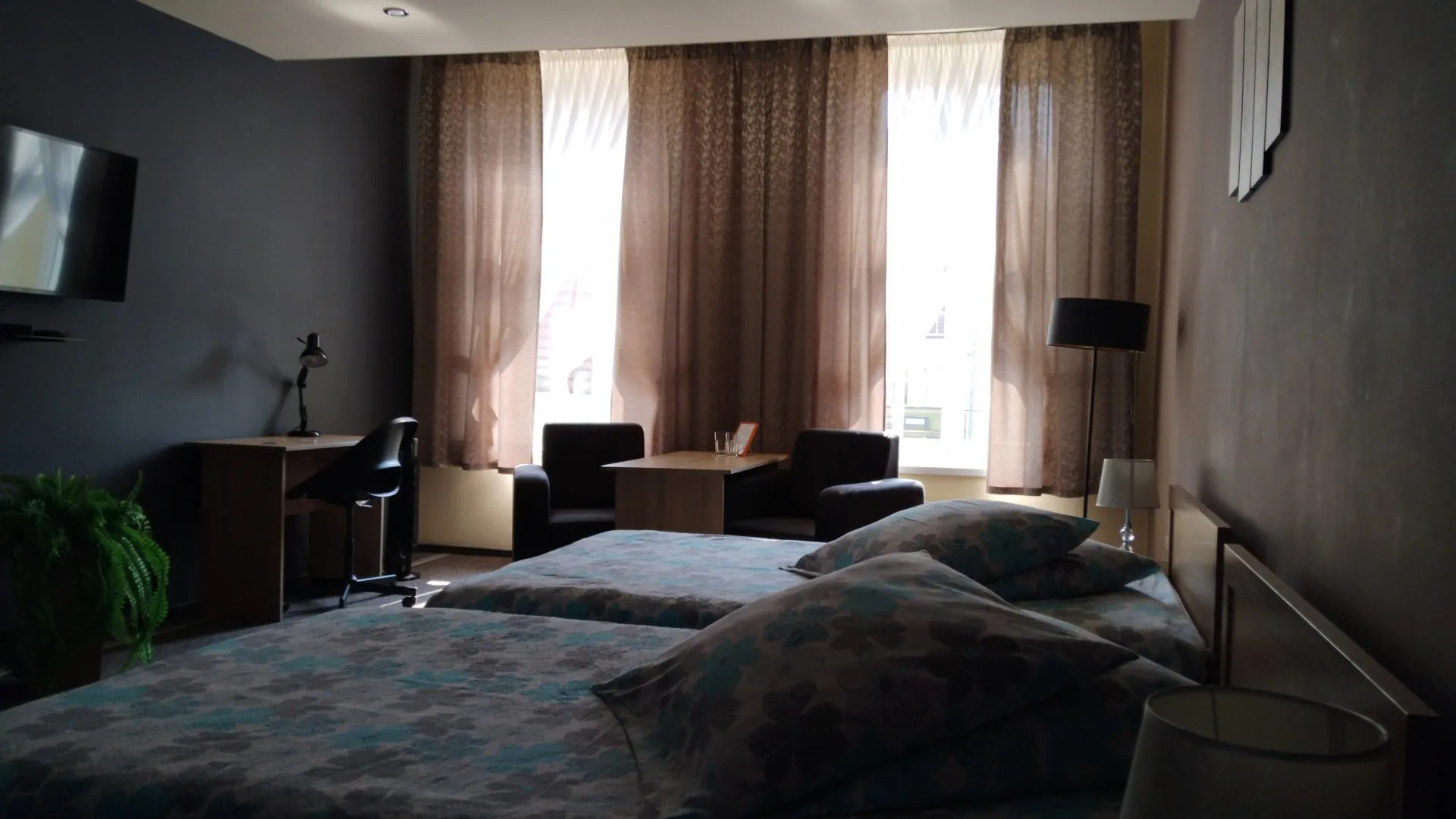 Przestronny pokój hotelowy z dwoma łóżkami pojedynczymi, biurkiem do pracy i wygodnym kącikiem wypoczynkowym przy oknie. Wnętrze utrzymane jest w stonowanych kolorach, z dużą ilością naturalnego światła wpadającego przez zasłonięte firany.