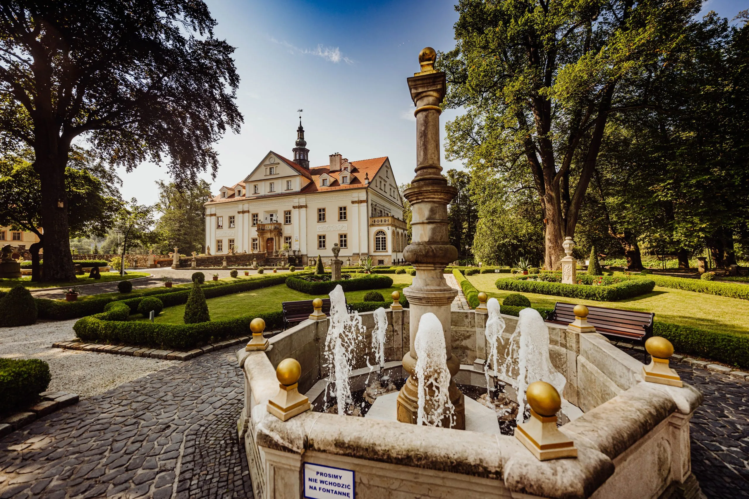 Zdjęcie przedstawiające Pałac Ciechanowice, ogród oraz czynna fontannę, którą widać na pierwszym planie. Zdjęcie wykonane przy słonecznej pogodzie.