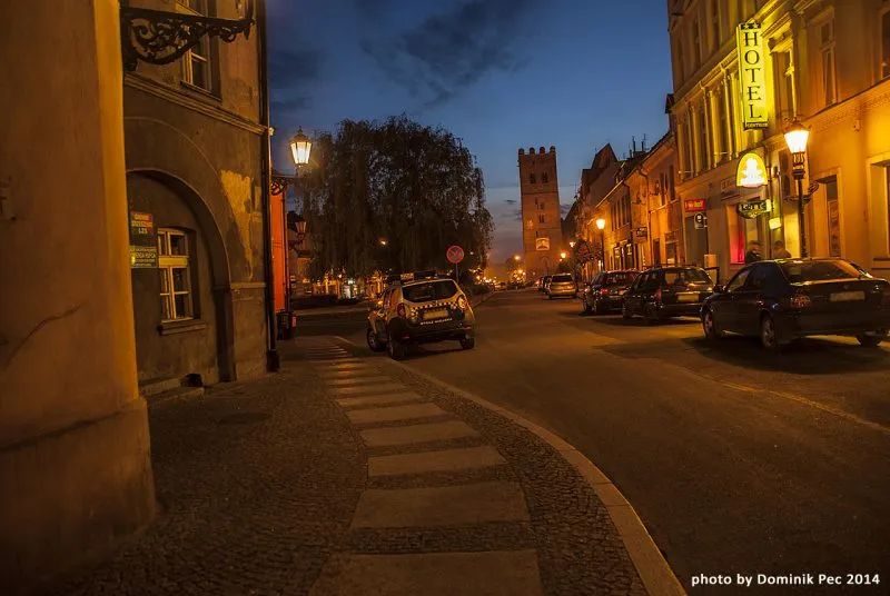 Wieczorny widok na ulicę w centrum miasta z charakterystyczną wieżą w tle. Po prawej stronie widać hotel z jasno podświetlonym szyldem, a po lewej staromiejską zabudowę.