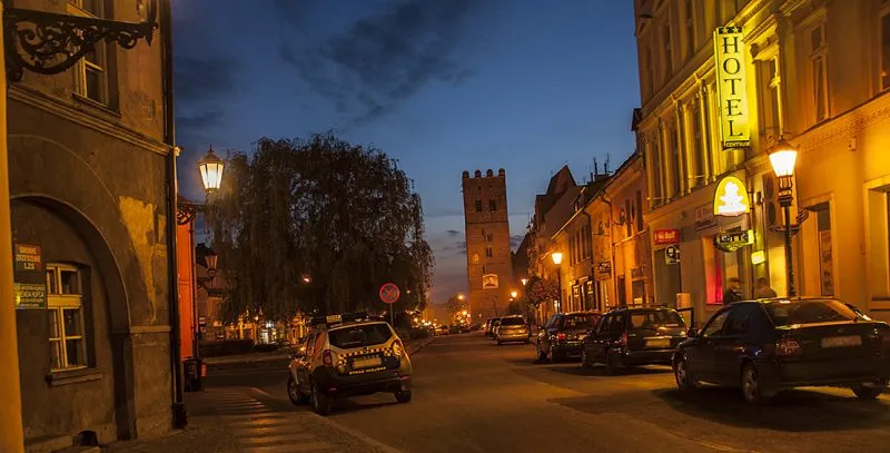 Wieczorny widok na ulicę w centrum miasta z charakterystyczną wieżą w tle. Po prawej stronie widać hotel z jasno podświetlonym szyldem, a po lewej staromiejską zabudowę.