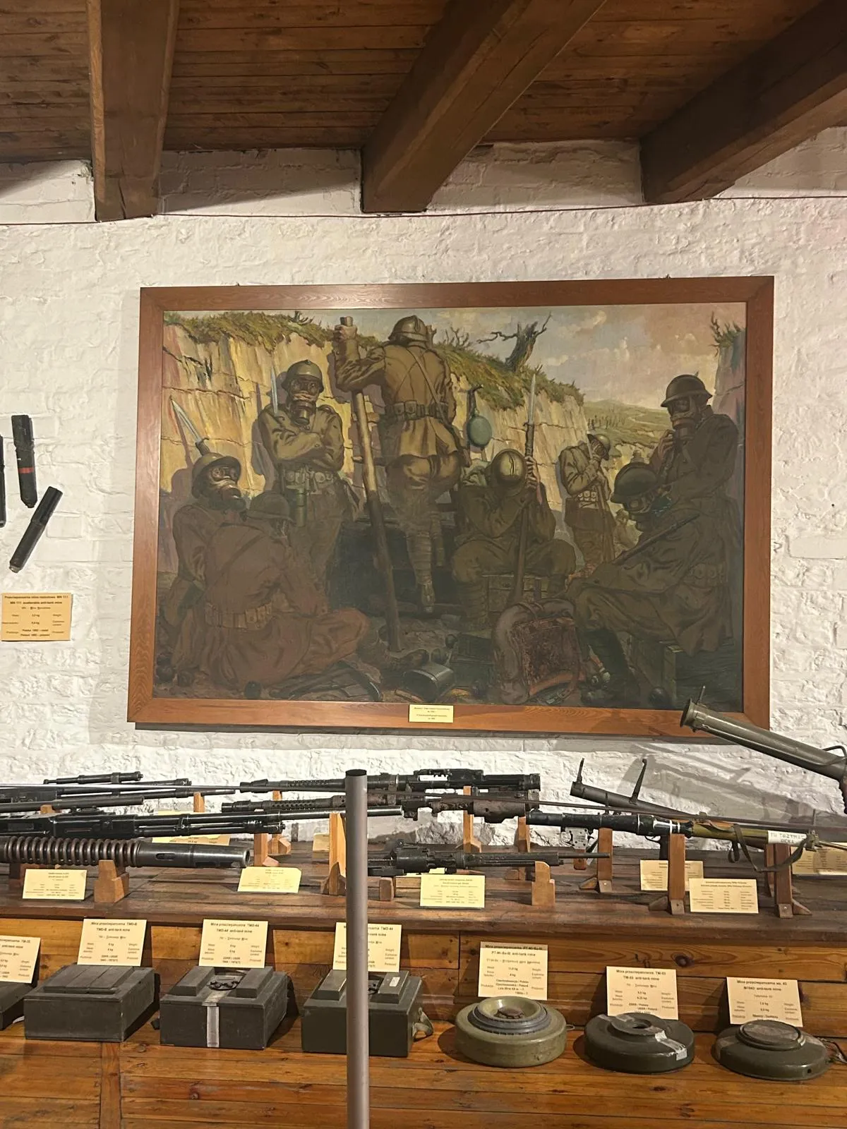 Zdjęcie przedstawia obraz żołnierzy znajdujących się w okopie. Pod obrazem ustawiona jest broń palna oraz miny