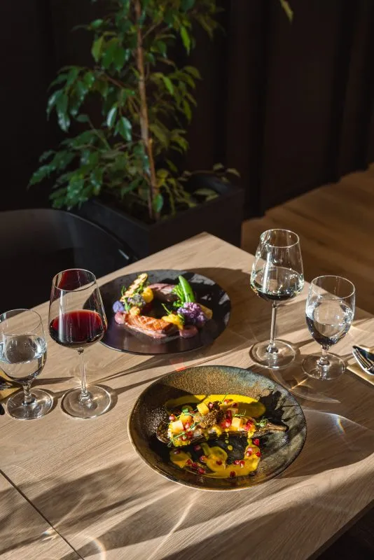 Na zdjęciu widać elegancko nakryty stolik w restauracji. Na stole ustawiono dwa wykwintnie podane dania oraz kieliszki z czerwonym winem i wodą. W tle widoczna jest doniczka z zieloną rośliną, a całość tworzy przytulną, nowoczesną atmosferę.