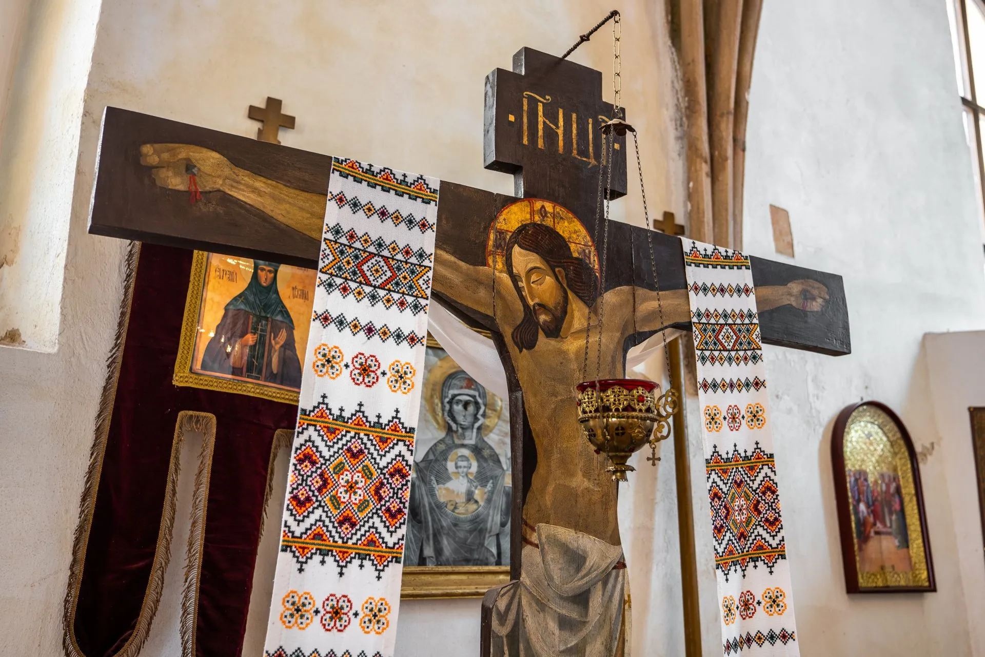 Drewniany krucyfiks w stylu bizantyjskim, z realistycznie przedstawioną postacią Chrystusa – smukłą, z wyraźnie zaznaczonymi ranami, głową pochyloną w lewo i złotą aureolą wokół głowy, zawieszony na tle ozdobnego ikonostasu.