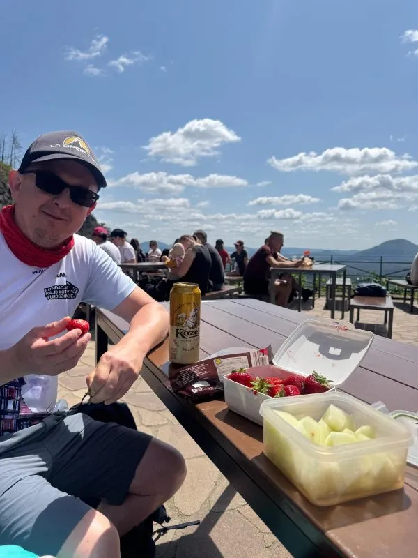 Rafał Mazurkiewicz sedí v čepici a slunečních brýlích na vyhlídkové terase a za ním jsou vidět turisté a rozsáhlé pohoří.