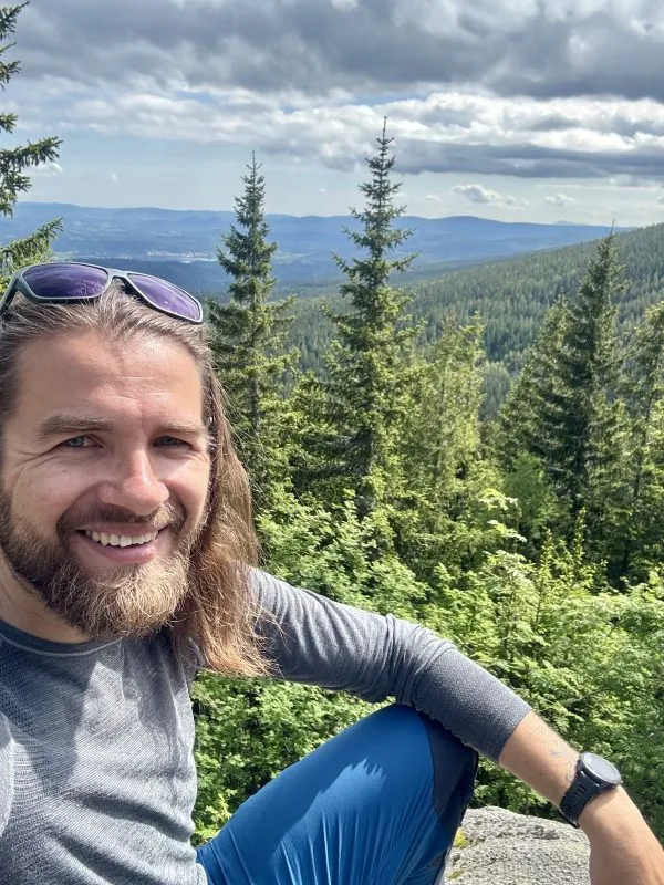 Michał Wawrzonek si dělá selfie na horské vyhlídce, sedí v šedém tričku před smrky a rozlehlými zalesněnými hřebeny.
