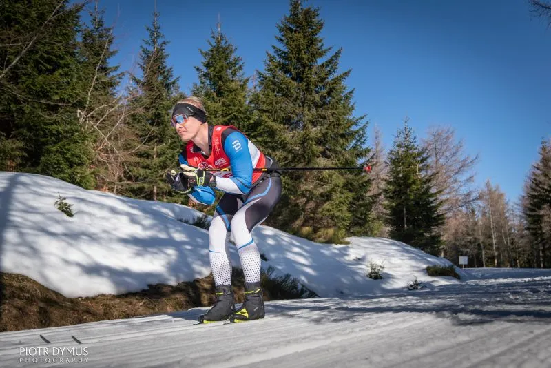 Uno sciatore di fondo in abbigliamento sportivo percorre un tratto primaverile del percorso innevato della Bieg Piastów, superando abeti sotto un cielo intensamente azzurro.