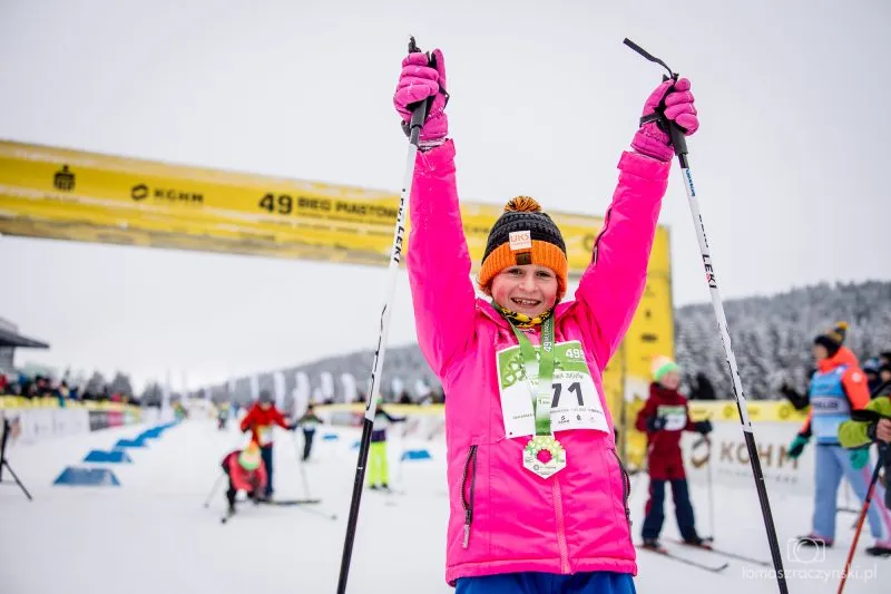 Un bambino con una tuta rosa solleva i bastoncini sopra la testa dopo aver completato la Bieg Piastów, in piedi sulla neve con una medaglia e il pettorale.