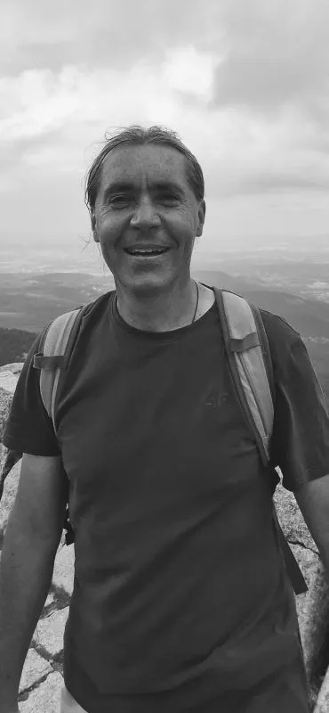 Lesław Wirbuł se usmívá na černobílém portrétu v turistickém tričku a s batohem, stojící na pozadí rozlehlé krajiny.