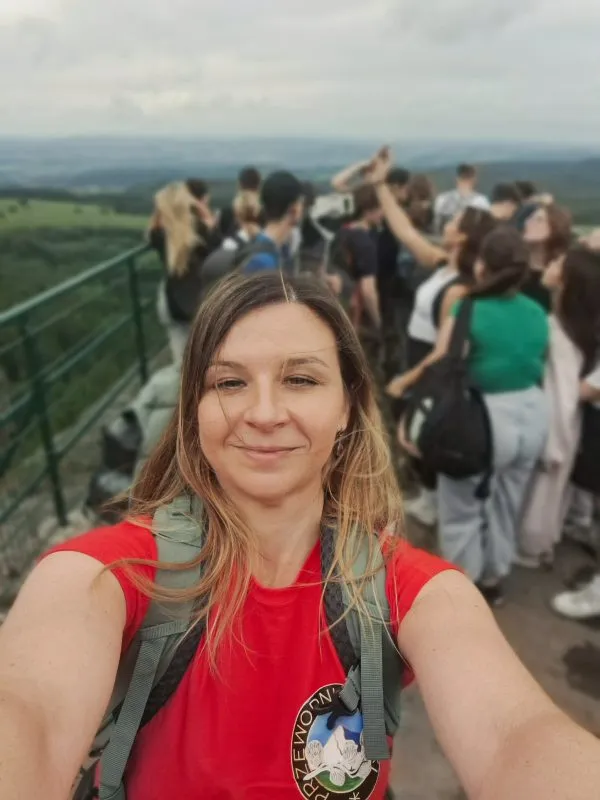 Anna Kopczak-Wirga si dělá selfie v červeném tričku na přeplněné vyhlídce s dřevěnou lávkou a zelenou horskou krajinou v pozadí.