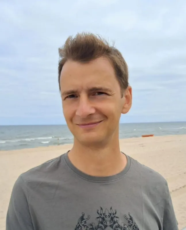 Przemek Lachowicz pózuje na písečné pláži v šedém tričku a za ním je vidět klidné moře, linie horizontu a lehce zamračená obloha.