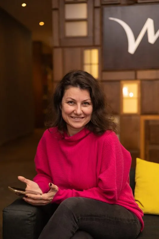 Tatiana Granowska sedí v růžové halence na černé pohovce během rozhovoru, gestikuluje rukama v teple osvětleném, elegantním interiéru.