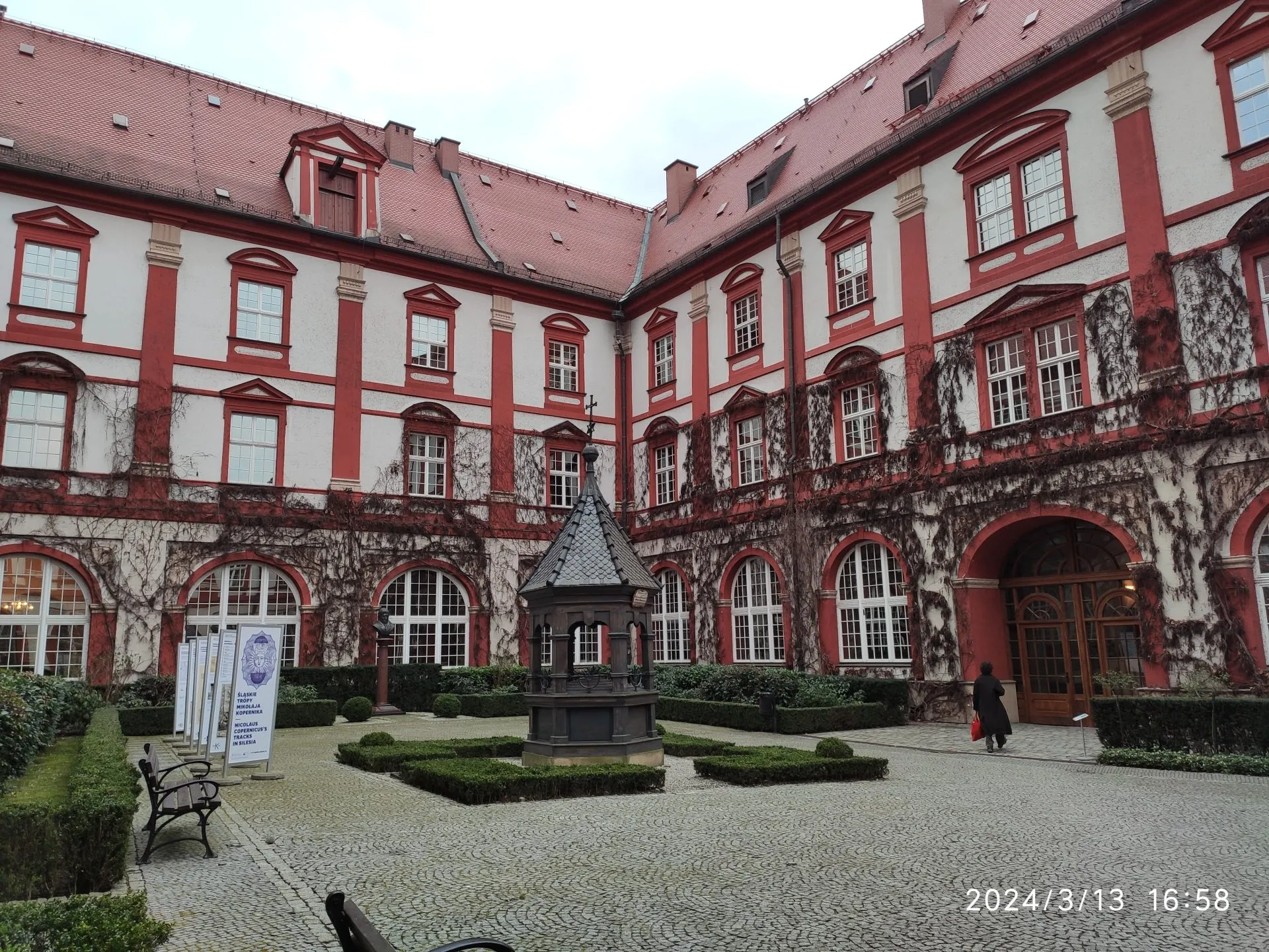 Dziedziniec zabytkowego budynku z czerwono-białą fasadą, ozdobioną suchym bluszczem i klasycznymi detalami architektonicznymi. Pośrodku znajduje się niewielka fontanna lub studnia, a całość otoczona jest przyciętymi krzewami i brukowaną nawierzchnią.
