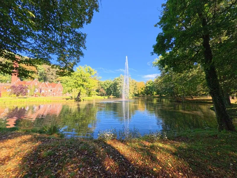 Lo stagno del parco di Kamieniec Ząbkowicki riflette la fontana, gli alberi e una parte del palazzo, mentre le rive ombreggiate sono ricoperte da prati e foglie autunnali.