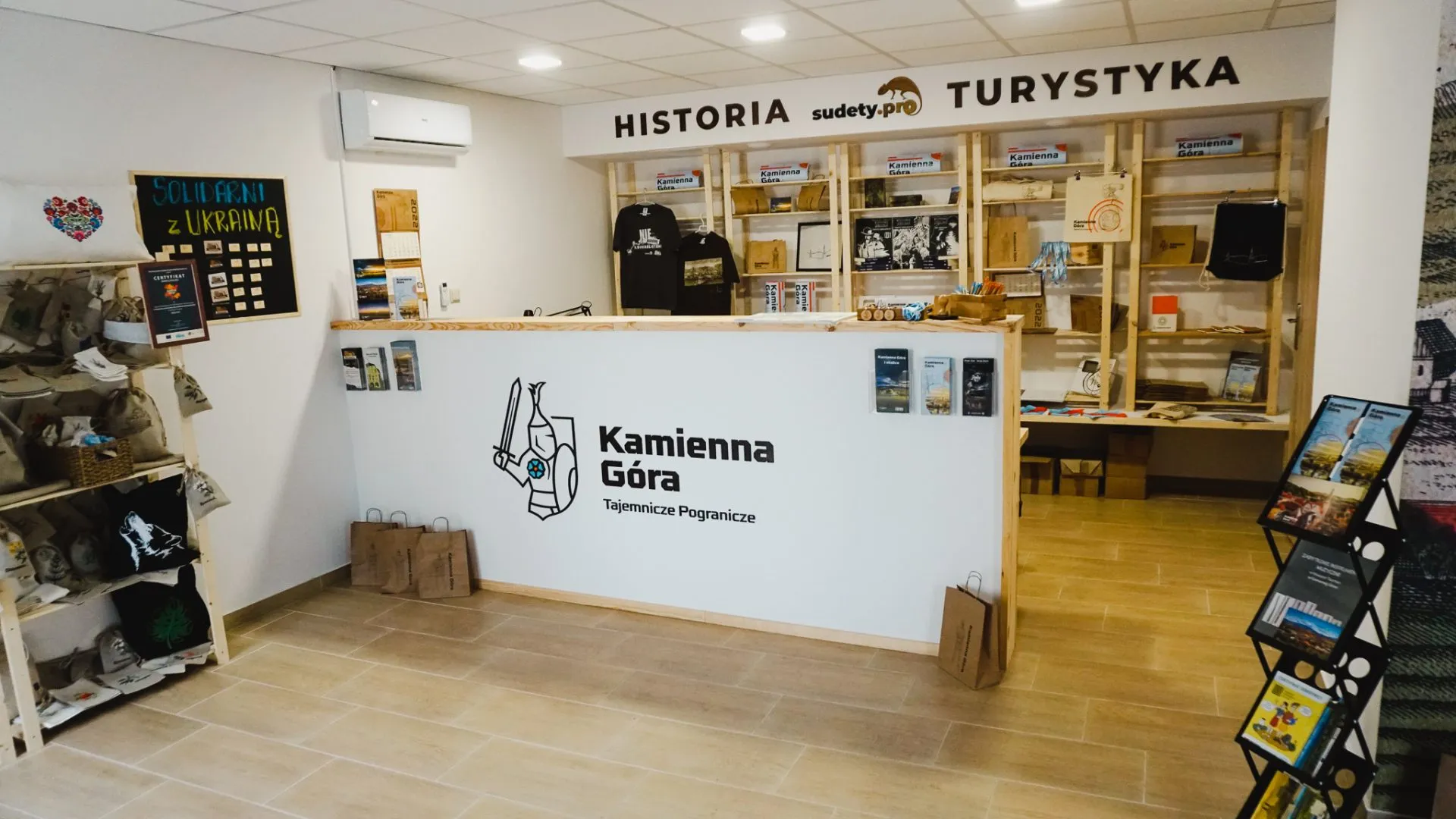 Bod sudetských turistických informací v Kamenné Hoře má dřevěný pult, police s průvodci a suvenýry a nástěnné nápisy „Historie“ a „Turistika“.