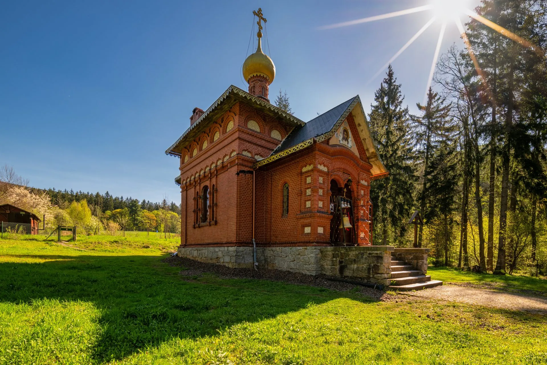 Na zdjęciu widać drewnianą cerkiew z kopulastymi dachami, otoczoną przez drzewa, z elegancką fasadą i prostą, ale dostojną architekturą, która emanuje spokojem i tradycją.