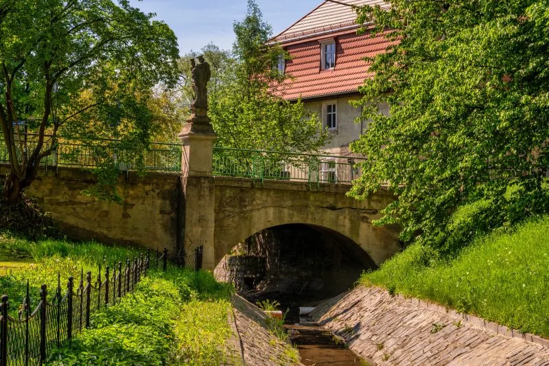 Na zdjęciu widać kamienny most z łukami, przechodzący nad rzeką, otoczony zielenią i zabudową miejską, z charakterystyczną XIX-wieczną architekturą.