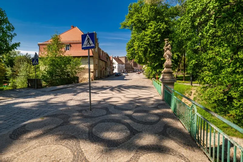 Na zdjęciu widać kamienny most z łukami, przechodzący nad rzeką, otoczony zielenią i zabudową miejską, z charakterystyczną XIX-wieczną architekturą.