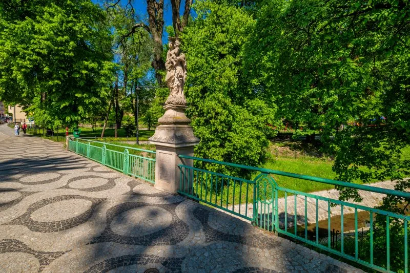 Na zdjęciu widać kamienny most z łukami, przechodzący nad rzeką, otoczony zielenią i zabudową miejską, z charakterystyczną XIX-wieczną architekturą.
