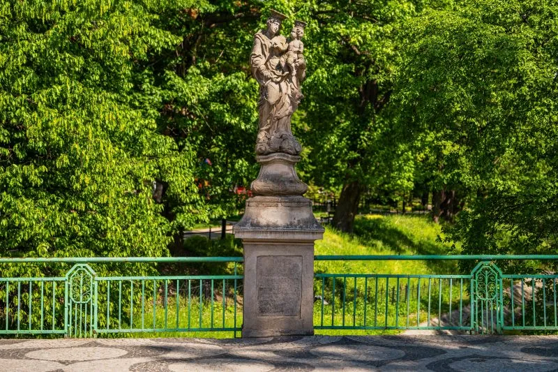 Na zdjęciu widać kamienny most z łukami, przechodzący nad rzeką, otoczony zielenią i zabudową miejską, z charakterystyczną XIX-wieczną architekturą.