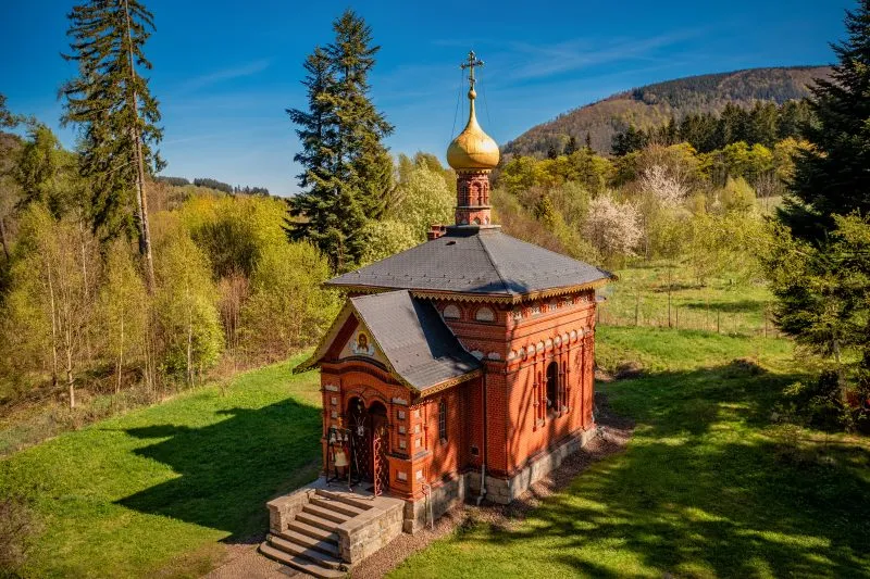 Na zdjęciu widać drewnianą cerkiew z kopulastymi dachami, otoczoną przez drzewa, z elegancką fasadą i prostą, ale dostojną architekturą, która emanuje spokojem i tradycją.
