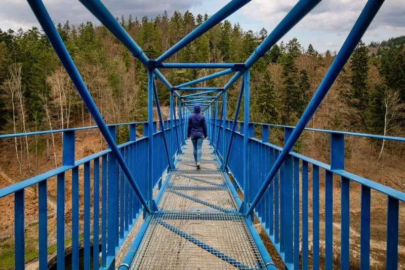 Modrá kovová lávka vede nad hlubokým údolím u přehrady v Międzygórze, obklopená strmými, hustě zalesněnými svahy.