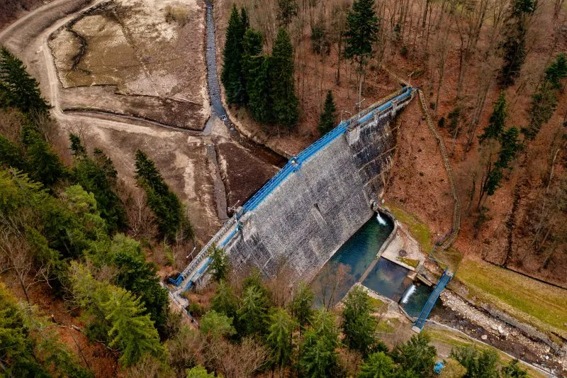 Pohled z ptačí perspektivy ukazuje kamennou přehradu v Międzygórze, její modrá zařízení a úzkou nádrž obklopenou strmými, zalesněnými svahy.