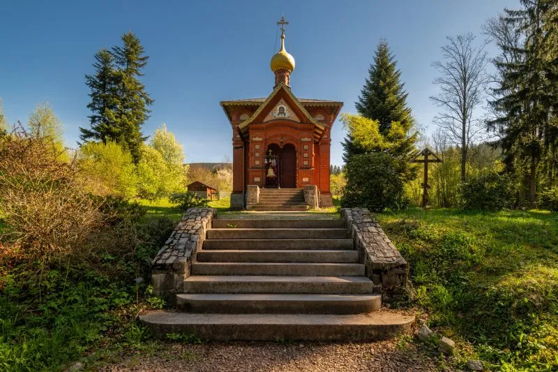 Na zdjęciu widać drewnianą cerkiew z kopulastymi dachami, otoczoną przez drzewa, z elegancką fasadą i prostą, ale dostojną architekturą, która emanuje spokojem i tradycją.