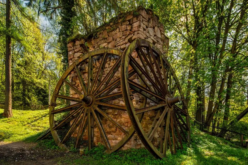 Zdjęcie pokazuje fragment starej, kamiennej konstrukcji z dwoma dużymi, drewnianymi kołami osadzonymi po bokach, otoczonej zielenią lasu.