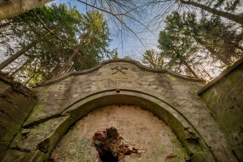 Na zdjęciu widać kamienny portal wejściowy sztolni Ernestine, otoczony przez zarośnięte wzgórze, z surowymi skałami i ścianą góry w tle.
