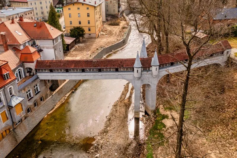 Kryty Most w Lądku-Zdroju to zabytkowa kamienna konstrukcja z łukowym przejściem, przykryta drewnianym dachem, otoczona bujną roślinnością i malowniczym krajobrazem.